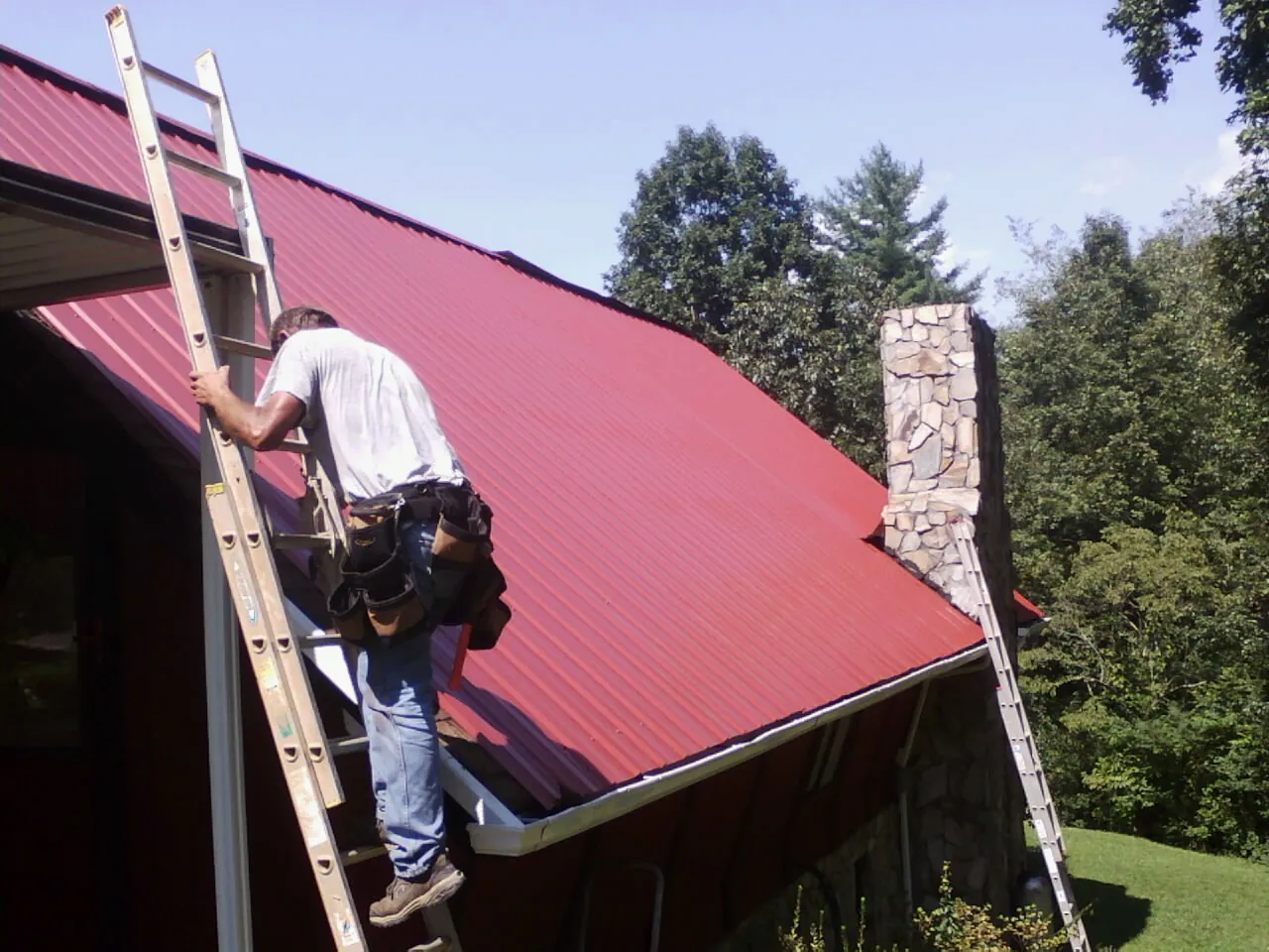 Roofer on a ladder beside a red metal roof and stone chimney — Soffit & Fascia Repair by Roofing Brothers in Quakertown